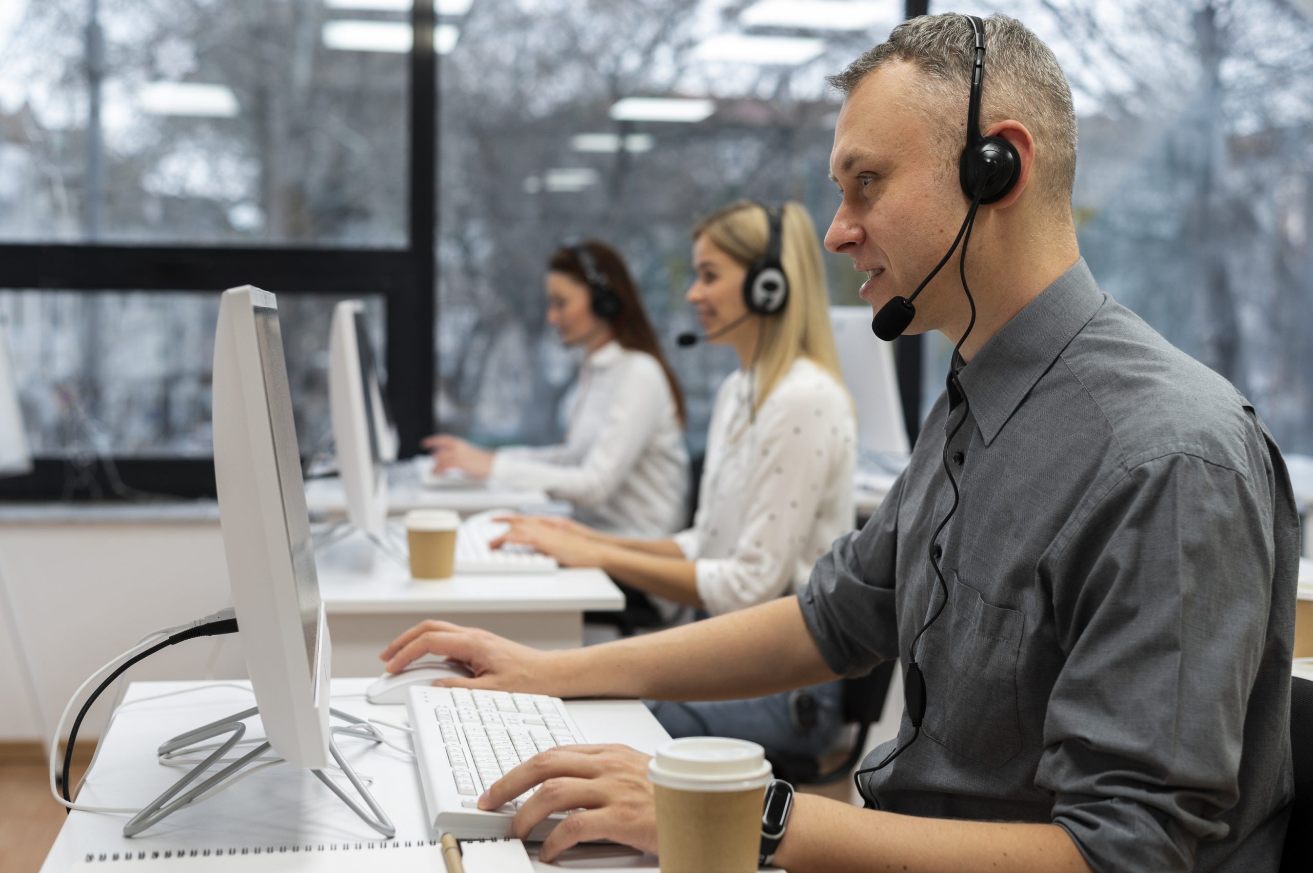 Na imagem temos 3 atendentes de telemarketing trabalhando. São 2 mulheres e 1 homem. Todos são brancos, usam headfone e estão sentados em mesas individuais de frente para computadores brancos. O homem, que aparece mais a frente na imagem é loiro de cabelos curtos, veste uma camisa cinza e na sua mesa tem um copo de café. Ao lado dele temos um mulher loira de cabelos longos que veste uma camisa branca e ao lado dela temos outra mulher de cabelos castanhos longos e que veste uma camisa branca.