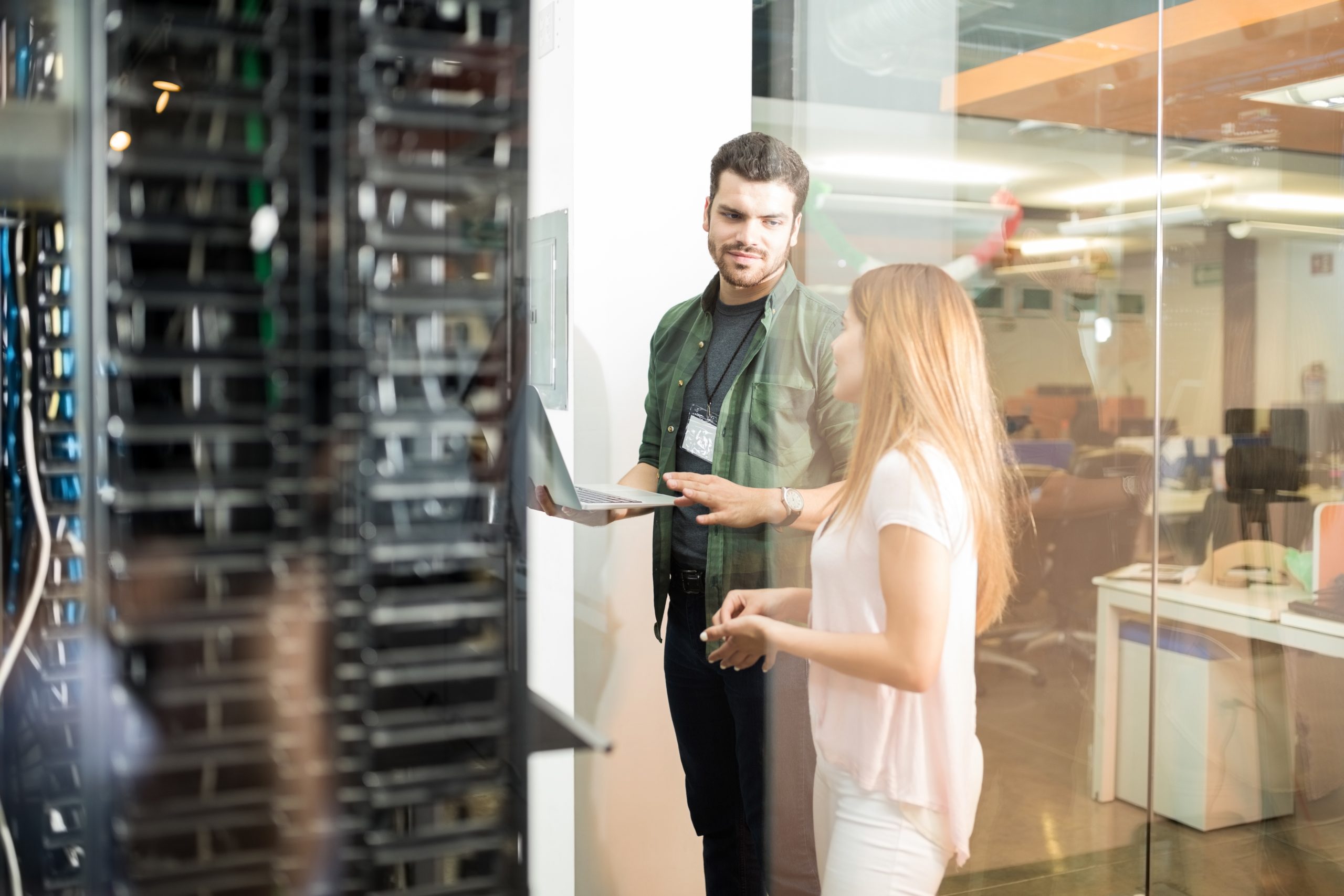 Na imagem temos um homem e uma mulher conversando em frente a um datacenter. O homem carrega nas mãos um notebook. Ele usa calça preta e camisa verde escuro e a mulher usa calça e blusa brancos.